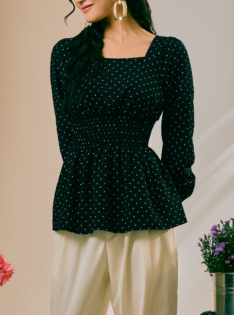 A woman wearing a dark green long-sleeve blouse with a white polka dot pattern and a square neckline, paired with cream-colored pants.