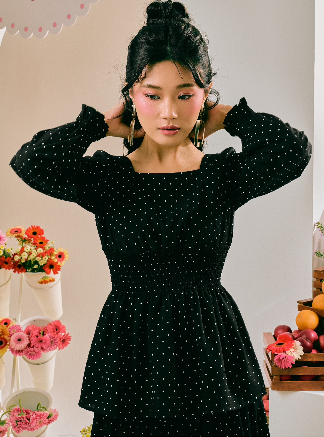 A woman wearing a black long-sleeve dress with a white polka dot pattern, standing in a decorated indoor setting with flowers and fruit visible in the background.