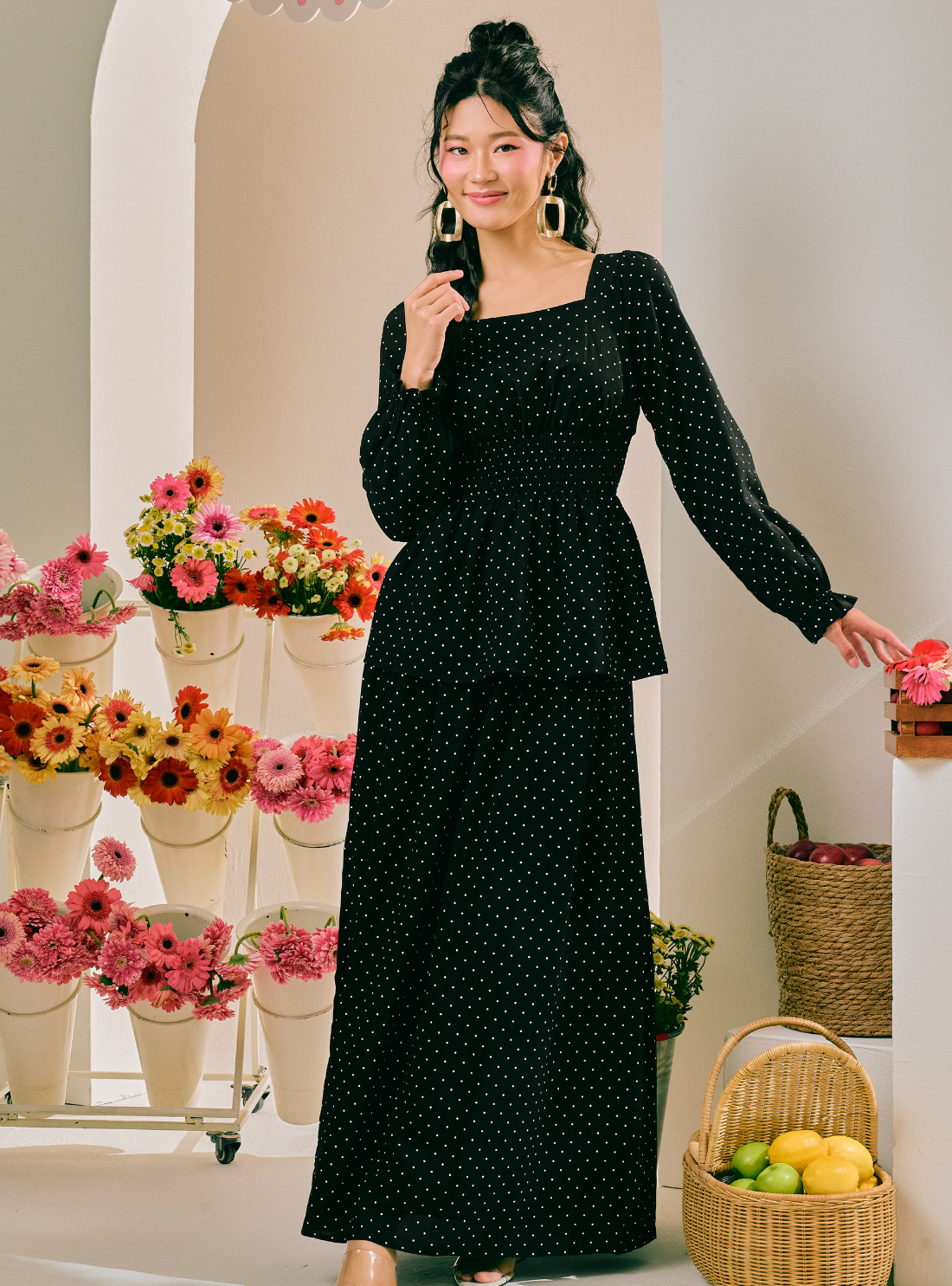 A woman models a black long-sleeve maxi dress with a small white polka dot pattern in a brightly lit indoor setting decorated with potted flowers and wicker baskets.