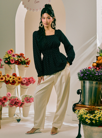 A woman models a black long-sleeve top with white polka dots and cream wide-leg pants in a styled studio setting with flower arrangements.