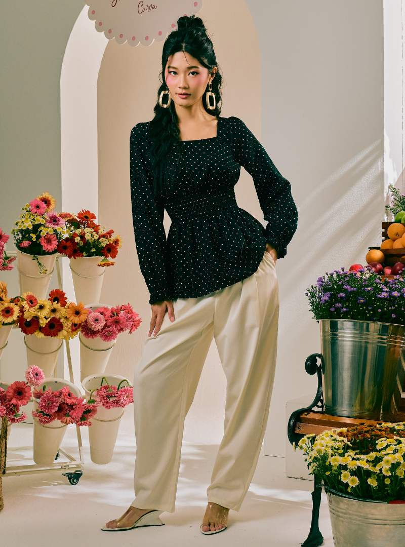 A woman models a black long-sleeve top with white polka dots and cream wide-leg pants in a styled studio setting with flower arrangements.