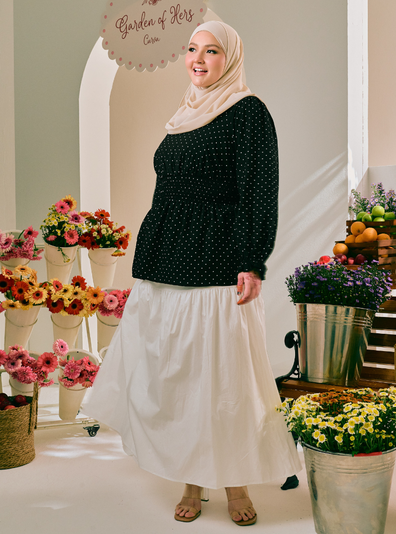 A woman wearing a black long-sleeve top with small white polka dots and a white maxi skirt, paired with a beige hijab, standing in a flower shop setting.