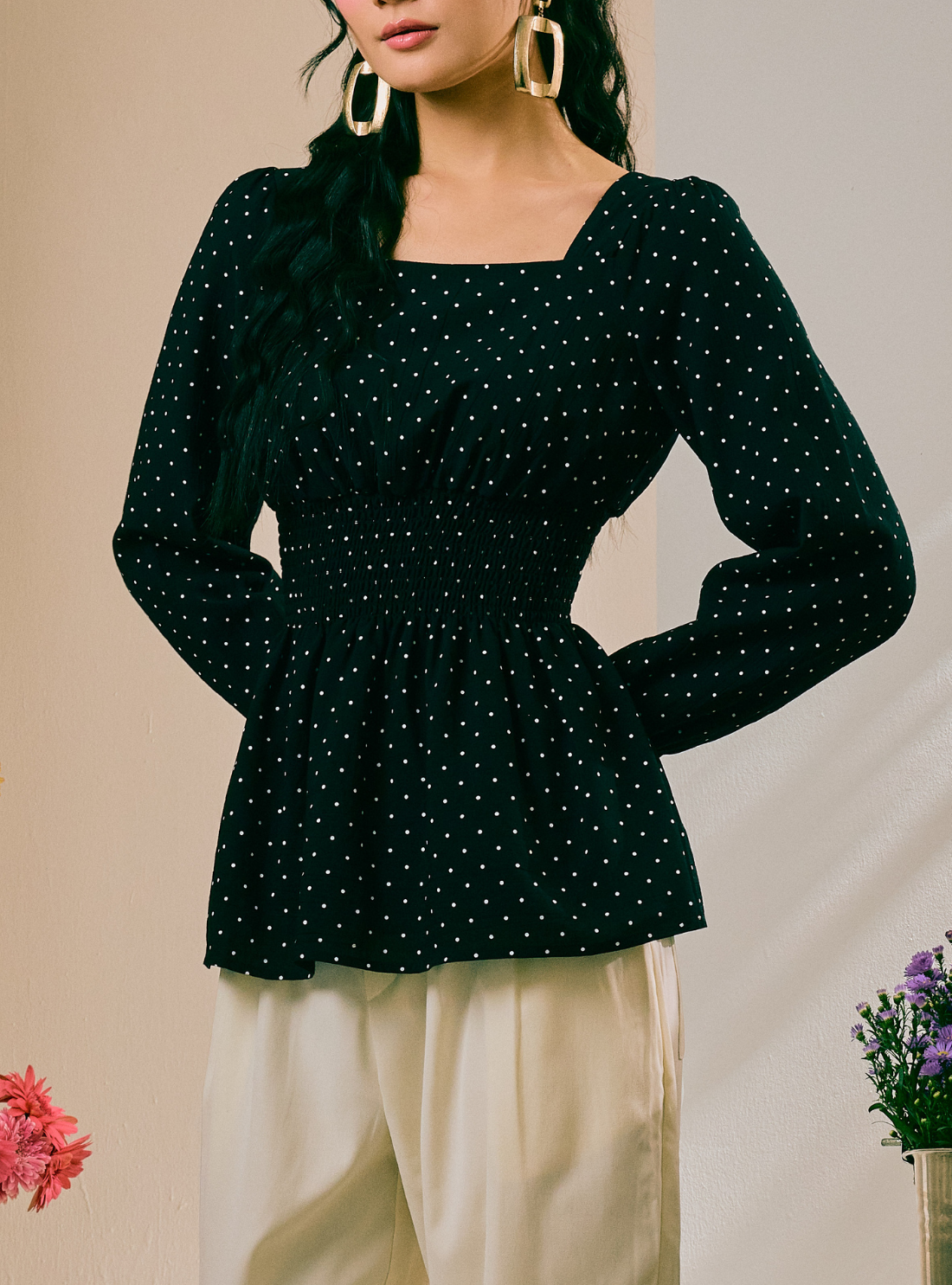 A woman wearing a dark green long-sleeve top with a white polka dot pattern and a square neckline, paired with cream-colored pants.