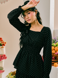 A woman wearing a black long-sleeve dress with a white polka dot pattern, featuring a square neckline and a peplum-style ruffled hem. She is posing in a brightly lit indoor setting with decorative flowers and fruit visible in the background.