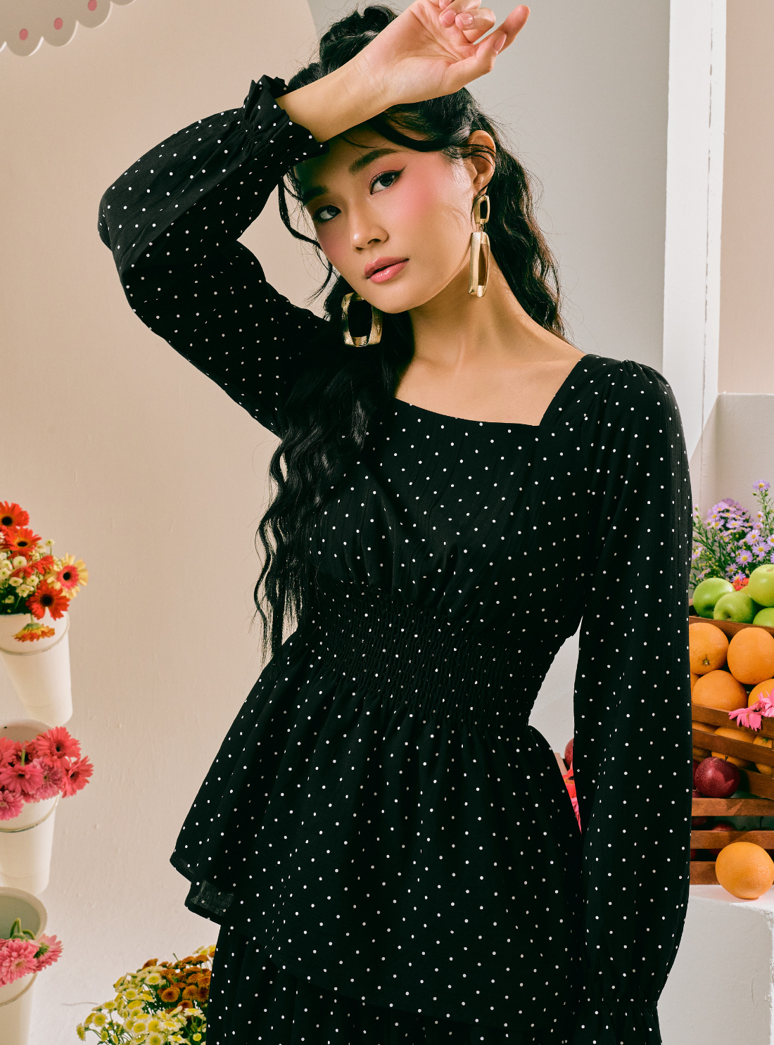 A woman wearing a black long-sleeve dress with a white polka dot pattern, featuring a square neckline and a peplum-style ruffled hem. She is posing in a brightly lit indoor setting with decorative flowers and fruit visible in the background.