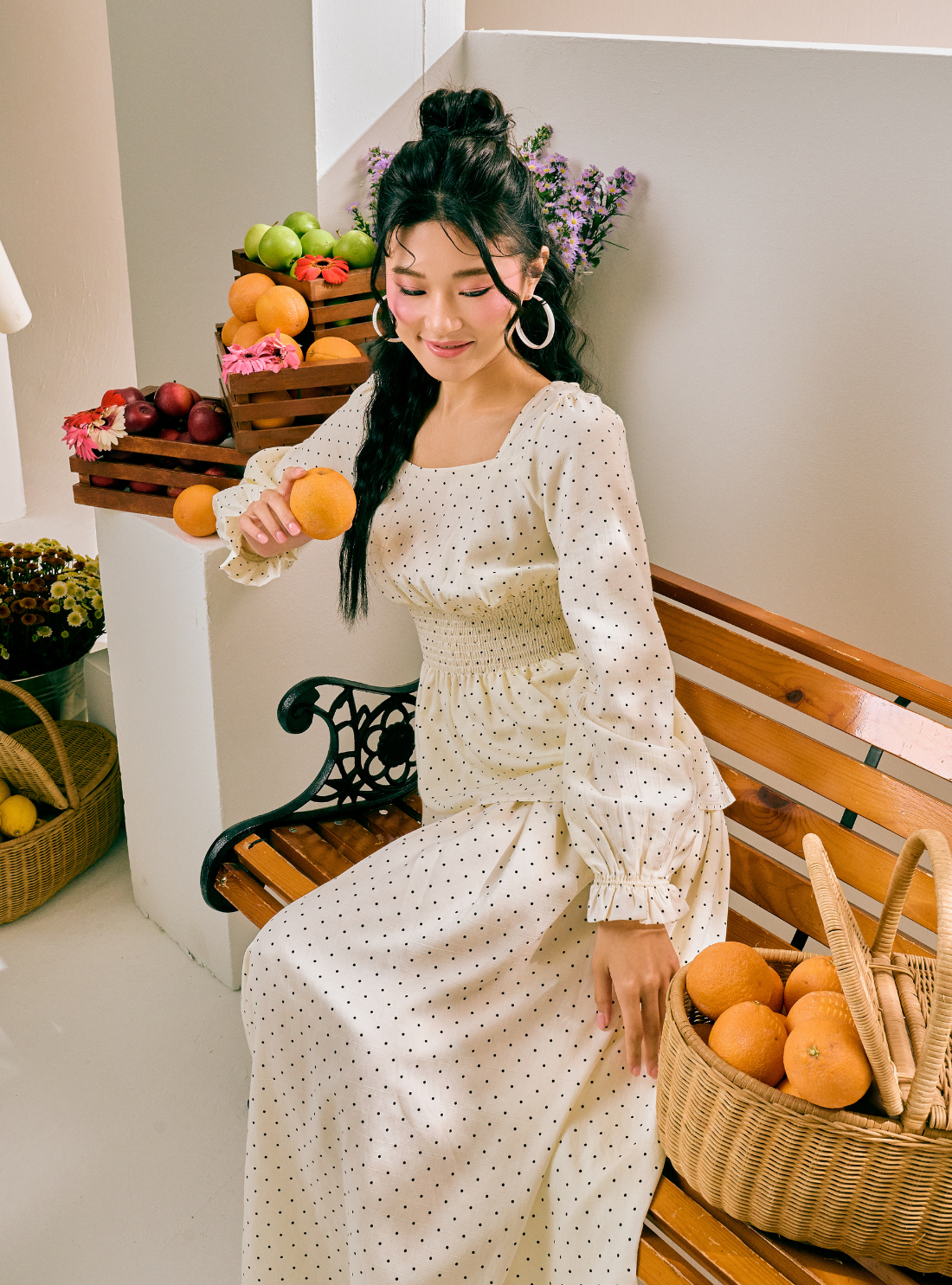 A woman sits on a wooden bench wearing a cream-colored, long-sleeved maxi dress with a small black polka dot pattern. The setting is decorated with baskets of oranges, flowers, and fruit crates against a neutral indoor background.