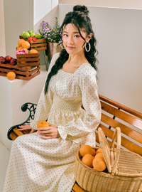 A woman sits on a wooden bench wearing a cream-colored long-sleeve dress with small black polka dots. She is holding an orange, with a wicker basket of oranges in the foreground and crates of fruit and flowers in the background.