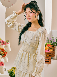 A woman wearing a cream-colored long-sleeve dress with a small black polka dot pattern and a smocked waist, posing in a brightly lit indoor setting decorated with flowers and fruit.