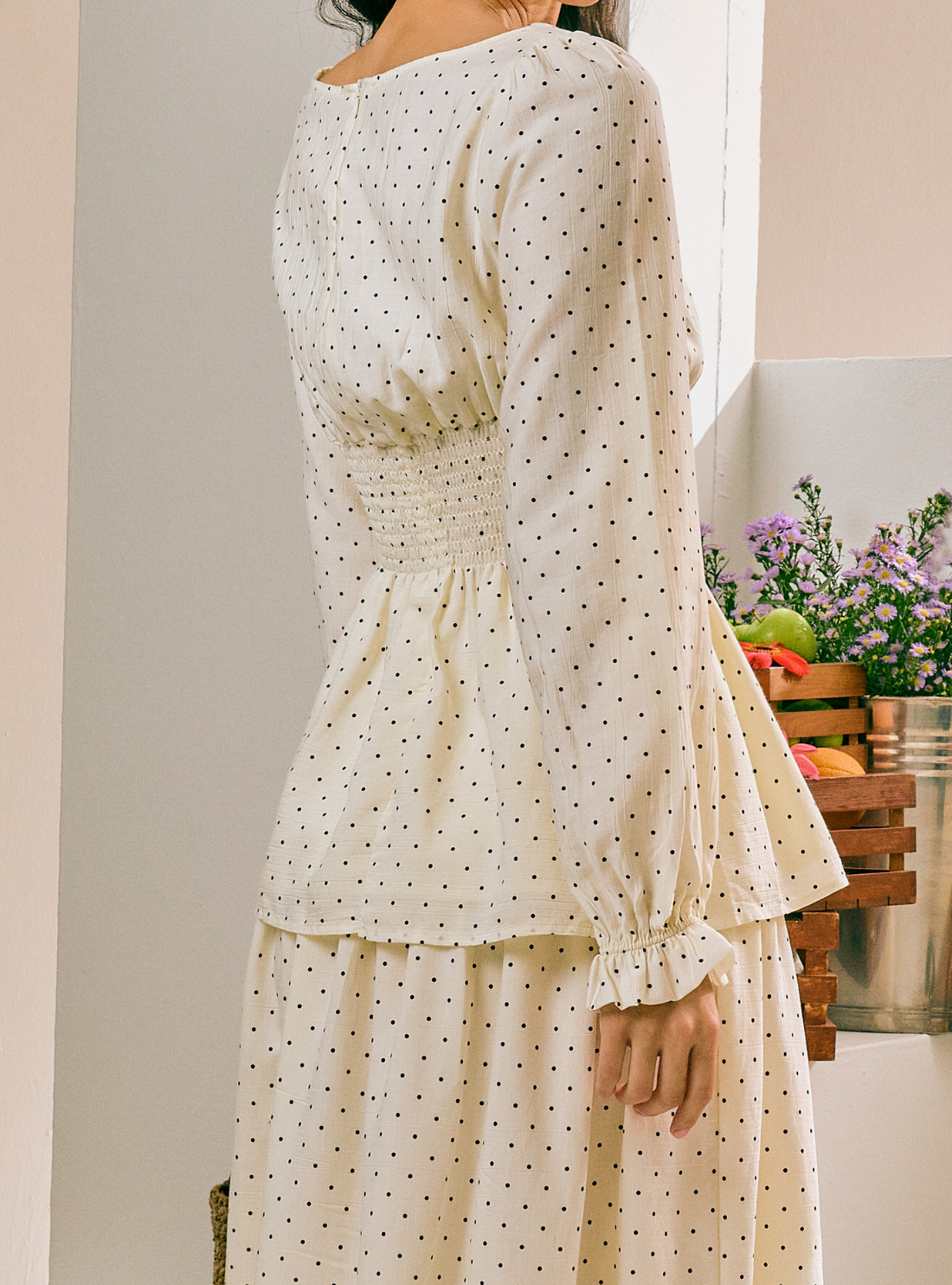 A woman wearing a cream-colored long-sleeve dress with a small black polka dot pattern, shown from a rear three-quarter angle in an indoor setting.
