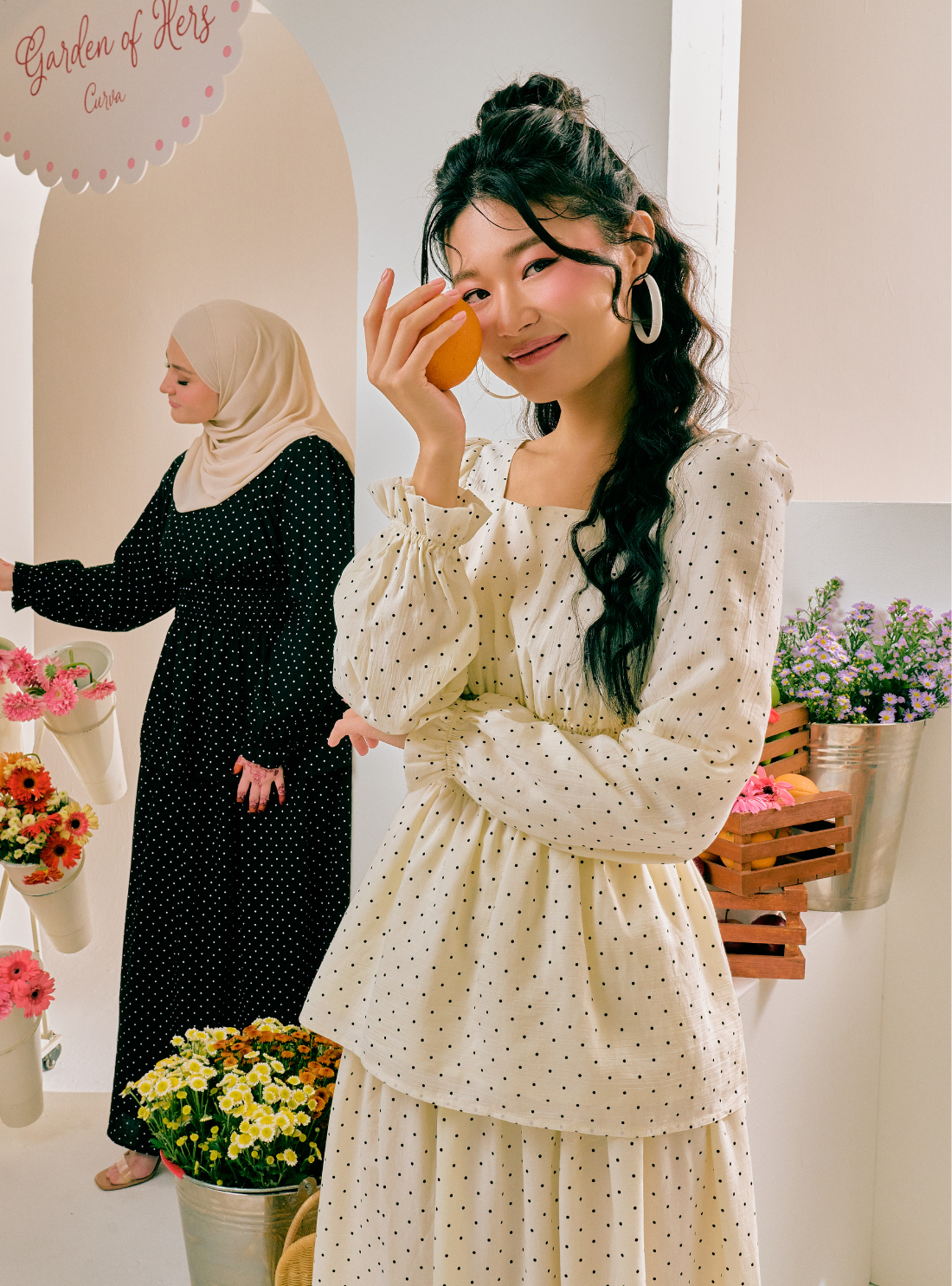 A woman in a cream and black polka dot long-sleeve dress holding an orange, with another woman in a black polka dot dress and beige hijab in the background of a boutique setting.