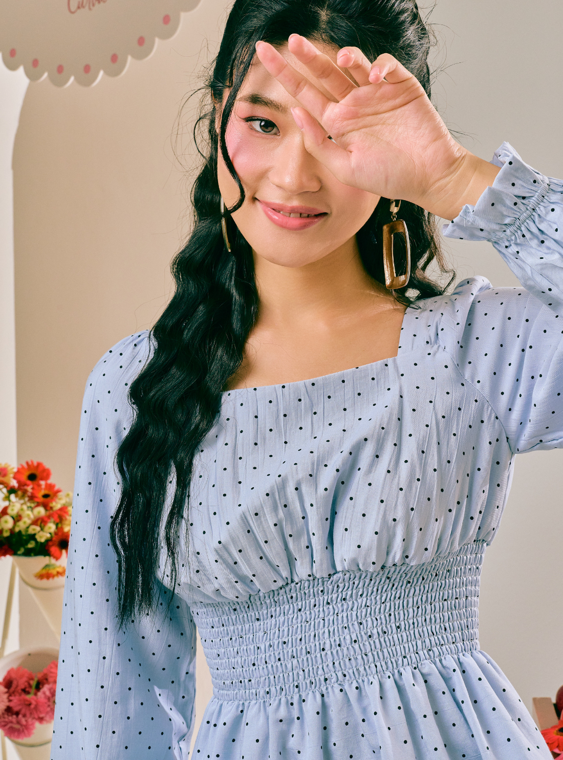 A woman wearing a light blue long-sleeve dress with a small black polka dot pattern, a square neckline, and a smocked waist. She is posing with one hand raised to shield her eyes, wearing gold hoop earrings.