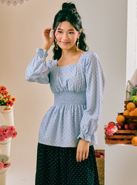 A woman modeling a light blue long-sleeve blouse with a small white polka dot pattern, a square neckline, and a smocked waist, paired with a dark green polka dot skirt, set against a warm beige background with decorative flowers and fruit.