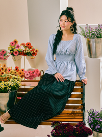 A woman sits on a wooden bench in a flower-filled room, wearing a light blue patterned long-sleeve blouse and a dark green polka-dot skirt.