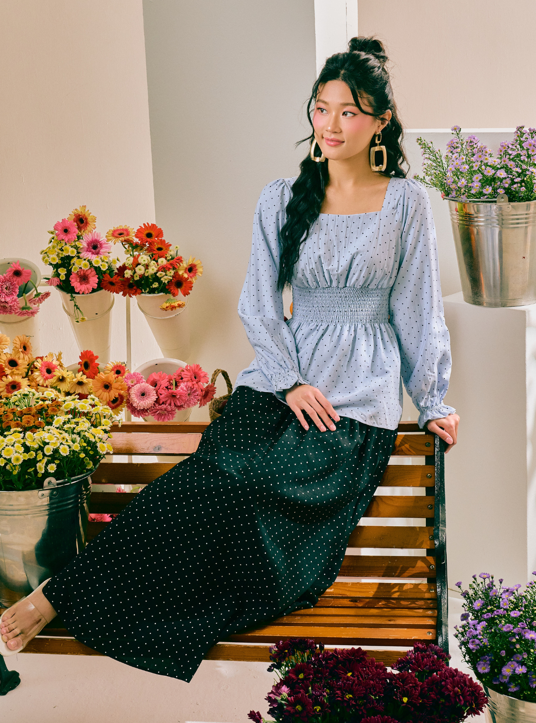 A woman sits on a wooden bench in a flower-filled room, wearing a light blue patterned long-sleeve blouse and a dark green polka-dot skirt.