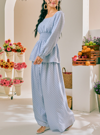 A woman models a light blue long-sleeve maxi dress with a small white polka dot pattern, featuring a smocked waist and a peplum-style hem, set against a bright, modern indoor studio backdrop with decorative flowers and fruit.