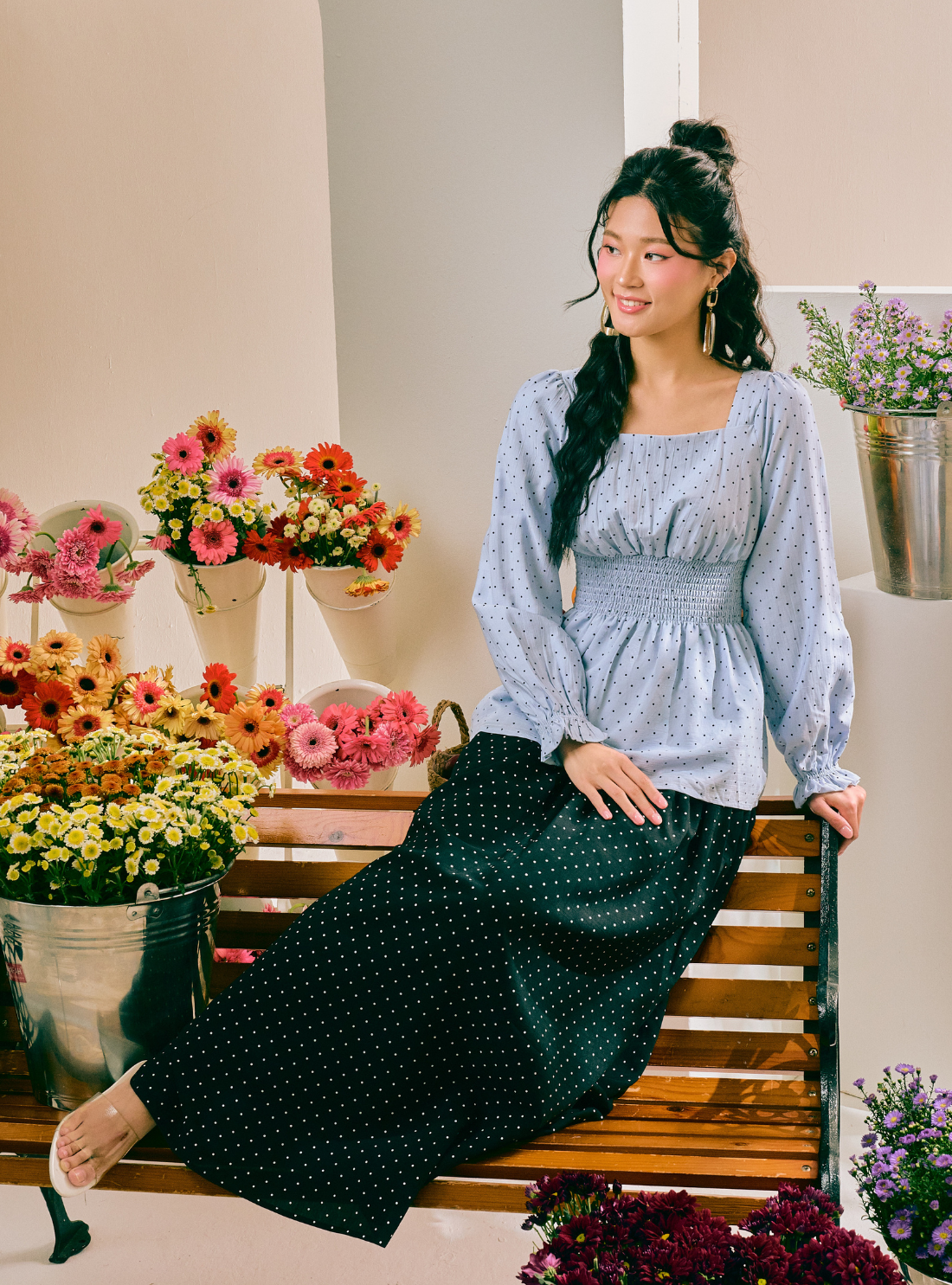 A woman sits on a wooden bench in a flower-filled room, wearing a light blue long-sleeve top with a small white pattern and a dark green wide-leg skirt with white polka dots.