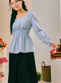 A woman models a light blue long-sleeve blouse with a small white polka dot pattern and a smocked waist, paired with a black polka dot skirt, in a styled indoor setting with flowers and baskets.