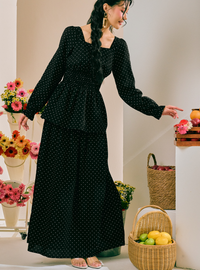 A woman wearing a long black dress with small white polka dots, standing in a styled indoor setting with flowers and woven baskets.