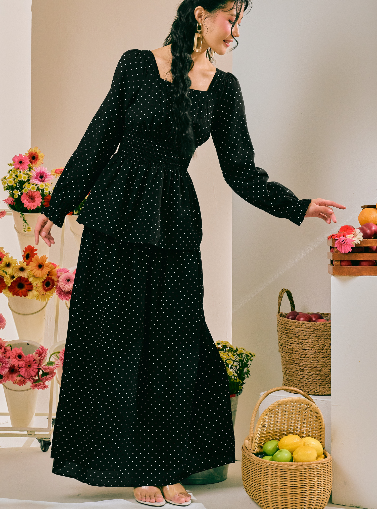 A woman wearing a long black dress with small white polka dots, standing in a styled indoor setting with flowers and woven baskets.