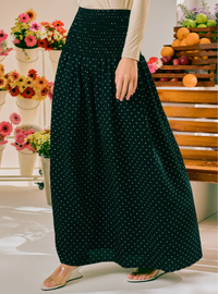 A dark green high-waisted maxi skirt with a small white polka dot pattern, worn by a model in a brightly lit indoor setting with flowers and fruit in the background.