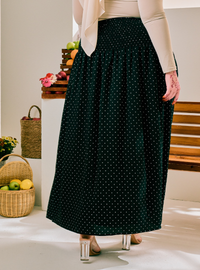 A person wearing a long dark green maxi skirt with small white polka dots, paired with a cream long-sleeve top and clear high heels, standing in a bright indoor setting with baskets and fruit in the background.