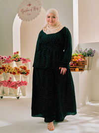 A woman wearing a dark green long-sleeve maxi dress with small white polka dots and a matching hijab stands in a bright indoor studio setting with flowers and fruit props.