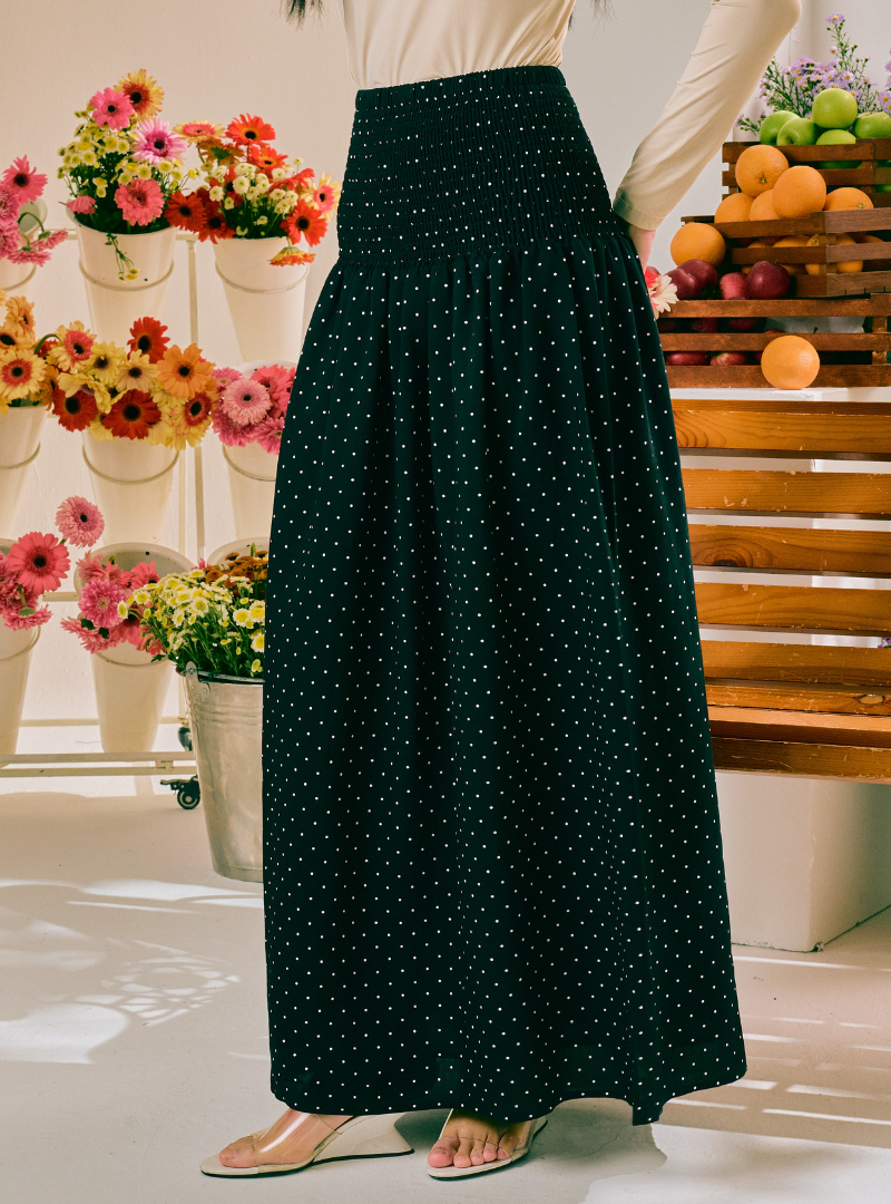 A dark green high-waisted maxi skirt with a white polka dot pattern, shown on a model in a styled indoor setting with flowers and fruit in the background.