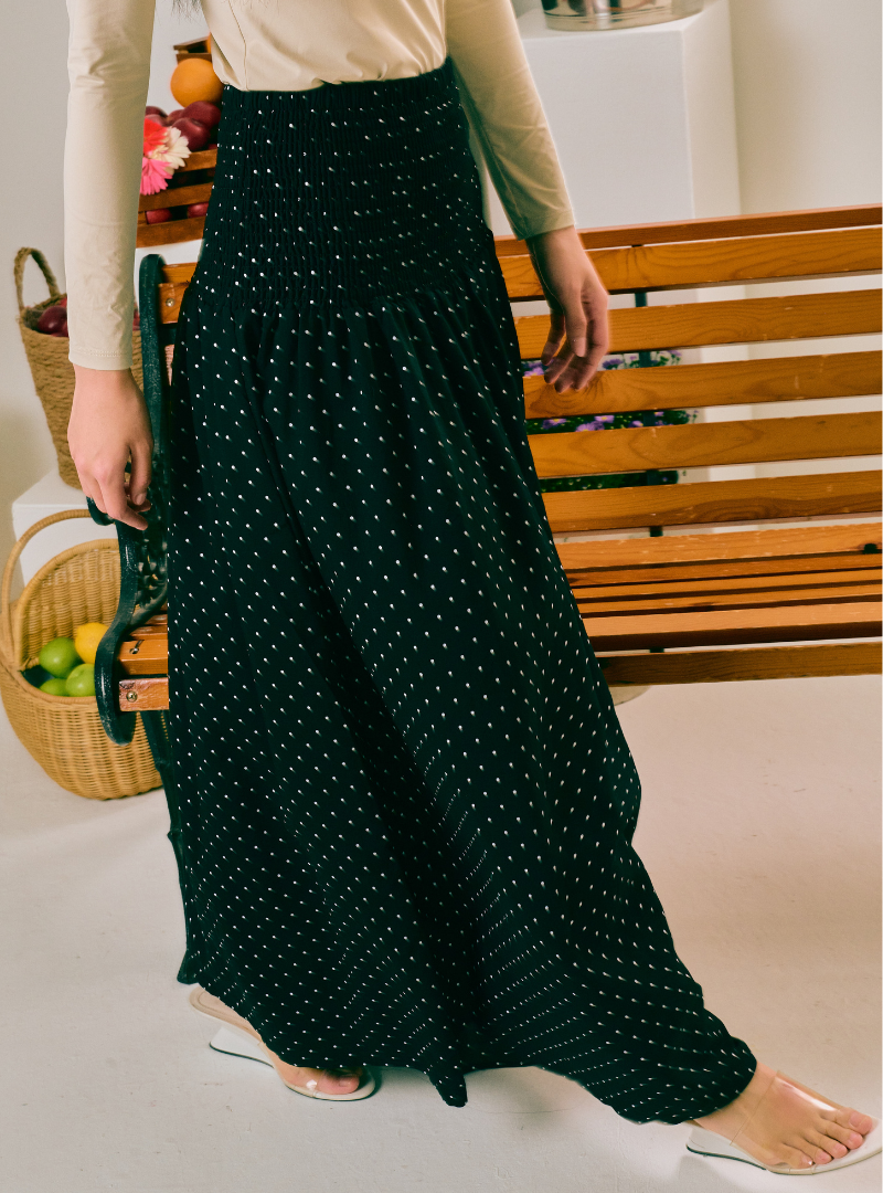 A person wearing a long black maxi skirt with a small white polka dot pattern, paired with a beige long-sleeve top and clear high-heel sandals, standing in front of a wooden bench.
