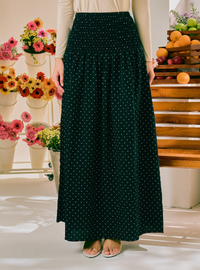 A woman wearing a long dark green maxi skirt with a white polka dot pattern, paired with a cream long-sleeve top and beige flat sandals. The background features a bright, clean studio setting with decorative flower arrangements on the left and a wooden crate of fruit on the right.