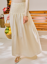 A woman wearing a cream-colored maxi skirt with a small black polka dot pattern and a smocked waistband, holding a wicker basket of fruit in a bright indoor setting.