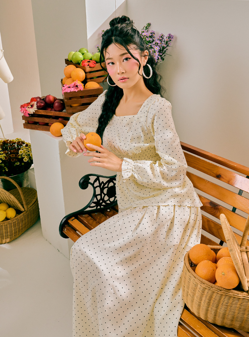 A woman sits on a wooden bench wearing a cream-colored, long-sleeved maxi dress with a small black polka dot pattern. She is holding oranges, with baskets of oranges and other fruits on the bench and beside her.