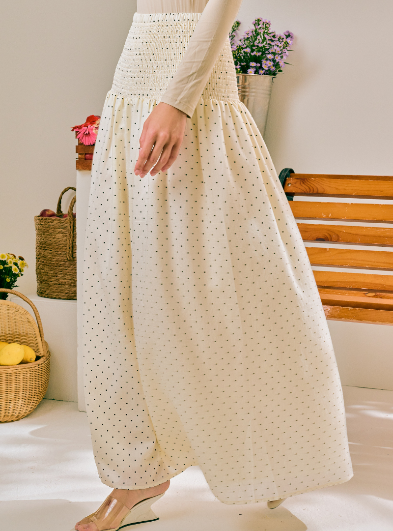 A person wearing a cream-colored, wide-leg maxi skirt with a small black polka dot pattern and a smocked waistband, paired with a matching cream long-sleeve top.