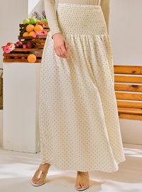 A woman wearing a long cream maxi skirt with small dark polka dots and a smocked waistband, paired with a beige long-sleeve top and clear heeled sandals. The background shows a white wall, a wooden bench, and stacked crates of fruit.