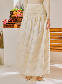 A woman wearing a long cream maxi skirt with a small black polka dot pattern and a smocked waistband, paired with a beige long-sleeve top and flat sandals. The background features a white wall, a wooden bench, and decorative wooden crates filled with fruit.