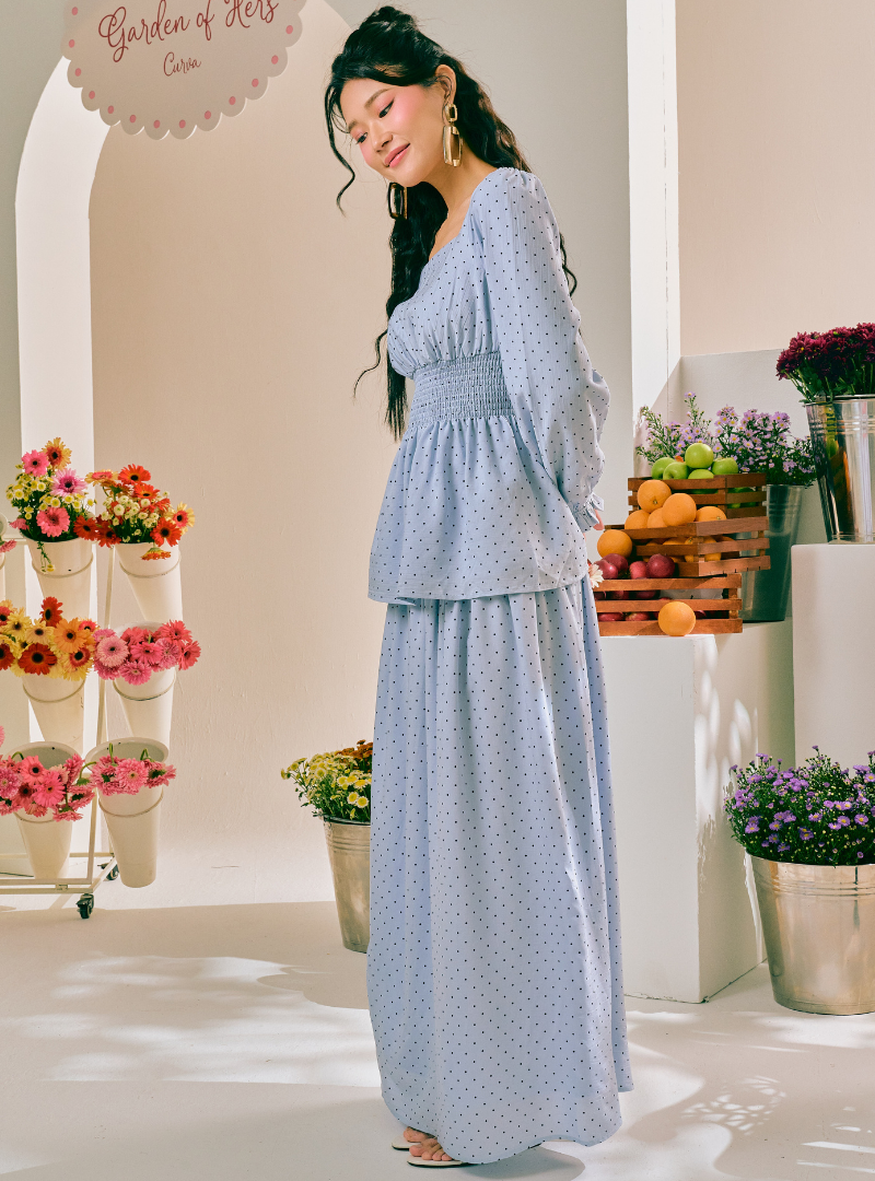 A woman modeling a light blue and white polka dot two-piece maxi dress with a peplum waist, set against a bright, sunlit studio backdrop decorated with flowers and fruit.
