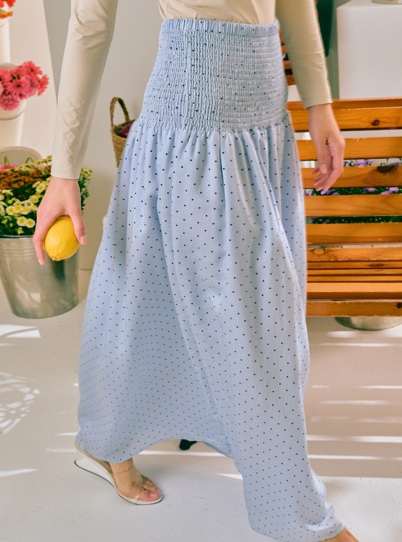 A person wearing a light blue high-waisted maxi skirt with a small black polka dot pattern and a smocked waistband, paired with a beige long-sleeve top and clear slide sandals.