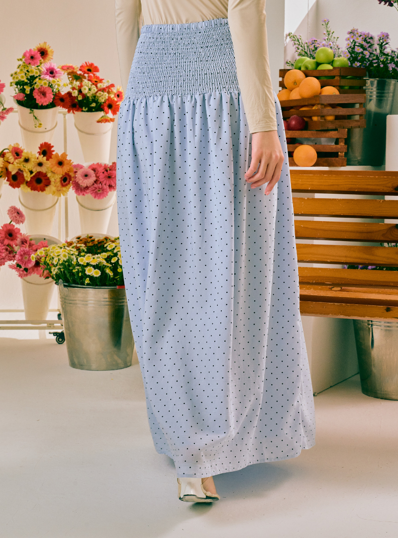 A person wearing a light blue maxi skirt with a small white polka dot pattern and a smocked waistband, styled with a cream long-sleeve top in a bright, flower-filled studio setting.