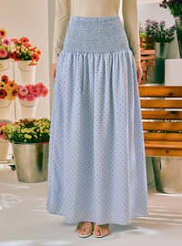 A light blue maxi skirt with a small black polka dot pattern and a smocked waistband, shown on a model in a brightly lit indoor setting with potted flowers in the background.