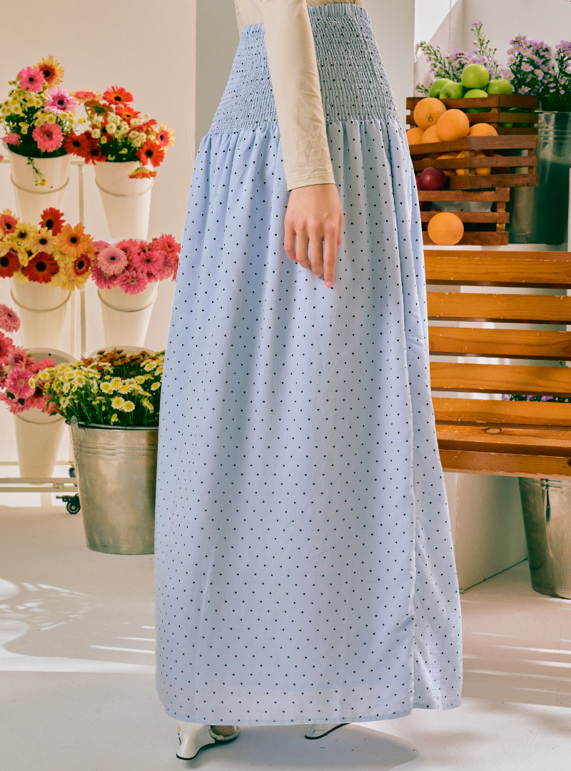 A light blue maxi skirt with a small white polka dot pattern and a smocked waistband, shown on a model in a brightly lit indoor setting with flowers and a wooden bench in the background.