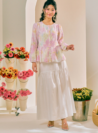 A woman models a two-piece outfit consisting of a light pink and white floral long-sleeve peplum blouse and a matching white tiered maxi skirt, set against a warm cream-colored studio background decorated with potted flowers.