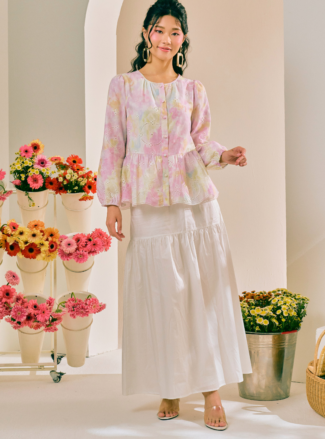 A woman models a two-piece outfit consisting of a light pink and white floral long-sleeve peplum blouse and a matching white tiered maxi skirt, set against a warm cream-colored studio background decorated with potted flowers.