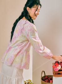 A woman wearing a light pink and cream eyelet embroidered long-sleeve blouse with a peplum waist, paired with a white skirt, standing next to a basket of fruit.