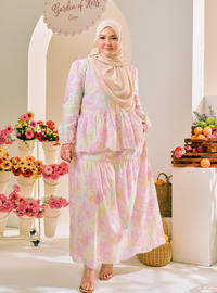 A woman wearing a long-sleeved, tiered modest maxi dress with a pastel floral print in pink, yellow, and green, paired with a beige hijab. She is standing in a bright, styled indoor setting with flower arrangements and fruit baskets.