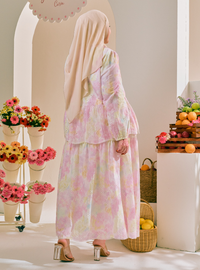 A woman wearing a light pink and white floral print two-piece modest outfit with a matching hijab, shown from behind in a bright, styled indoor setting with flowers and fruit.