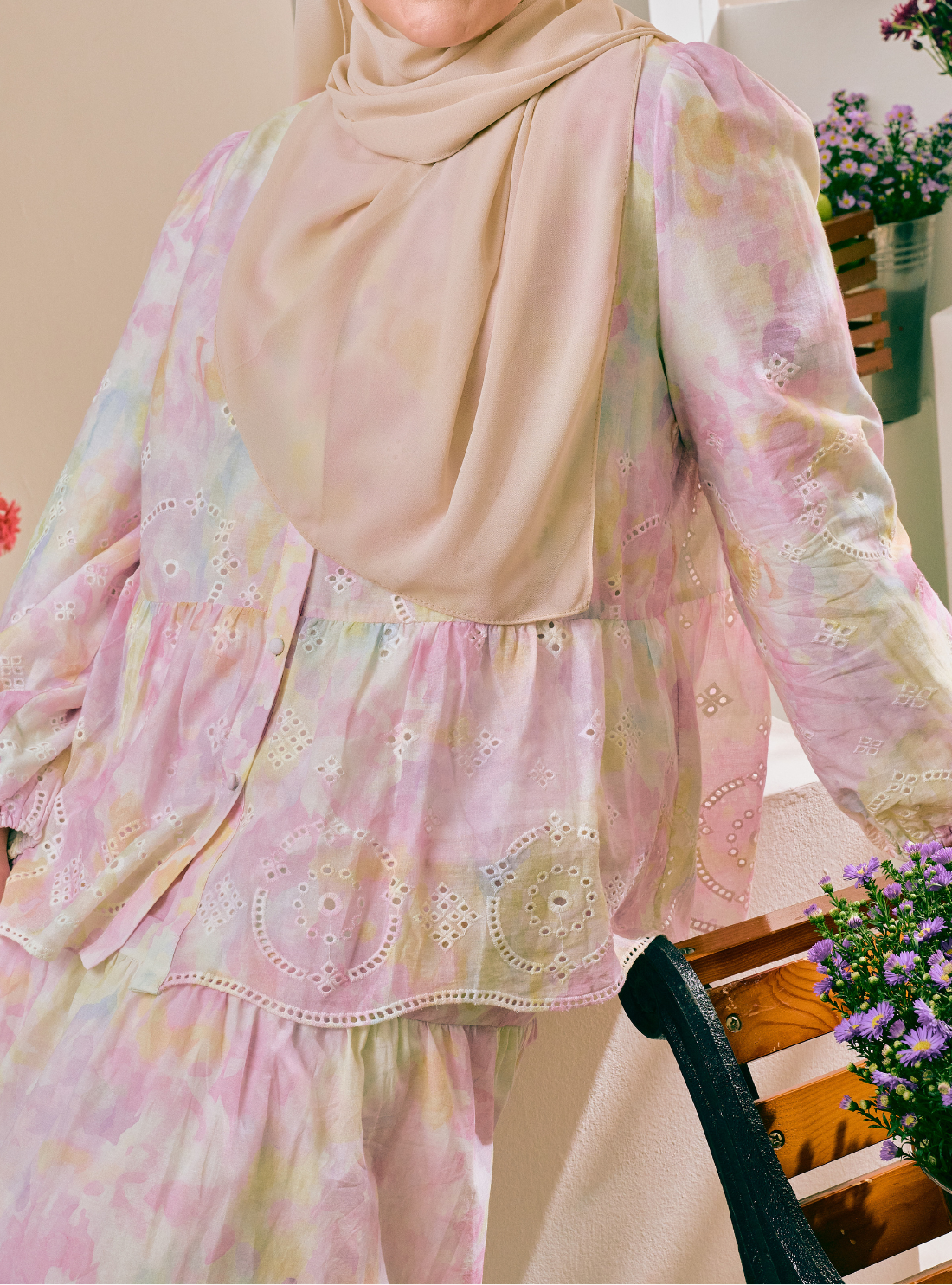 A close-up of a person wearing a pastel tie-dye traditional outfit with a matching beige hijab. The outfit features a long-sleeved tunic top with eyelet embroidery and a tiered skirt, in soft pink, yellow, and green tones.