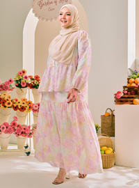 A woman wearing a light beige hijab and a long-sleeved, tiered maxi dress with a pastel pink, yellow, and green watercolor floral print. She is standing in a bright, decorated studio setting with flowers and fruit baskets.