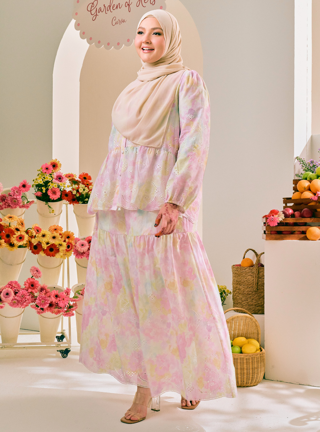 A woman wearing a light beige hijab and a long-sleeved, tiered maxi dress with a pastel pink, yellow, and green watercolor floral print. She is standing in a bright, decorated studio setting with flowers and fruit baskets.