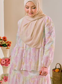 A woman wearing a light beige hijab and a long-sleeve tiered blouse with a pastel multicolor floral print, standing in an indoor setting with decorative flowers and a basket in the background.