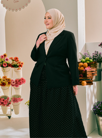 A woman wearing a beige hijab, a black pinstripe blazer, and a black midi skirt with white polka dots, standing in a brightly lit indoor setting decorated with flowers and fruit.