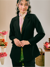 A woman wearing a black pinstriped blazer over a light pink scarf and green top, standing in a studio setting with decorative flowers and a basket.